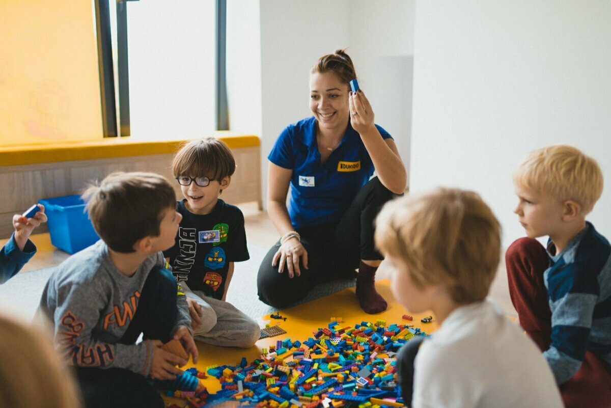 A group of children sitting on the floor playing with colorful LEGO bricks. Sitting with them is an adult, smiling and dressed in a blue shirt, holding a LEGO piece. The room is bright, with sunlight streaming in through the large windows - perfect for photo coverage of events in Warsaw.  