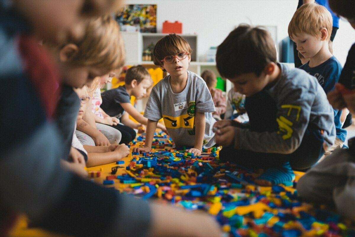 A group of young children sit on the floor and play with colorful Legos. One child wearing glasses and a gray shirt with a yellow pattern stands in the middle, focused on the blocks around them. The background shows the arrangement of the classroom, beautifully captured by event photographer Warsaw.  