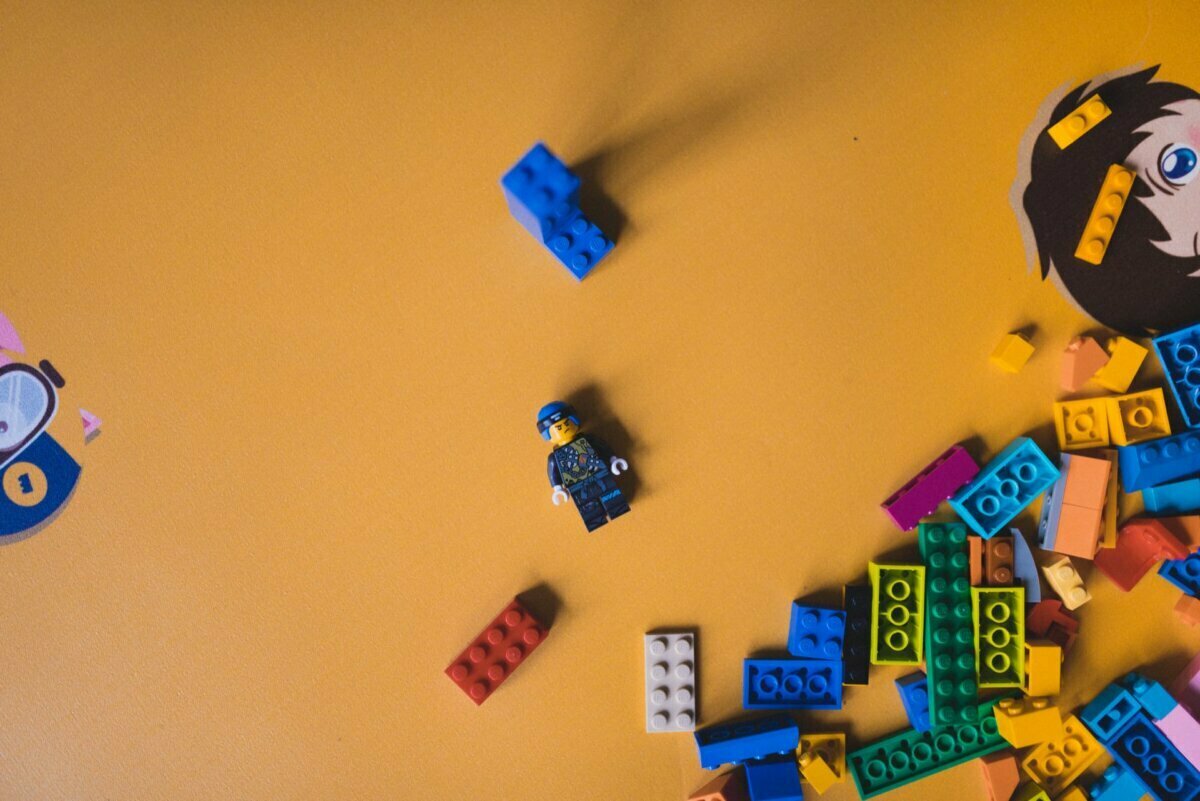 A colorful assortment of LEGO bricks is scattered on a yellow surface. Near the center is a LEGO minifigure dressed as a blue-clad worker. To the right, part of a cartoon face is visible, and a small blue LEGO piece stands vertically above the figure - perfect for photo opportunities of events.  