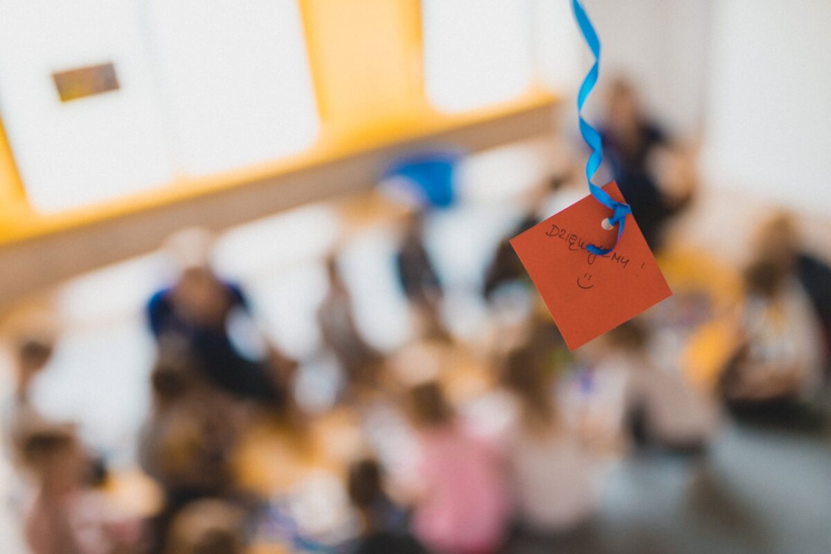 A red note with the words "Thank you!" (Thank you in Polish), and in the foreground a smiley face hangs on a blue ribbon. In the background is a group of people, mostly children, gathered in a blurry environment, probably a classroom or activity space - a perfect shot for event photography.