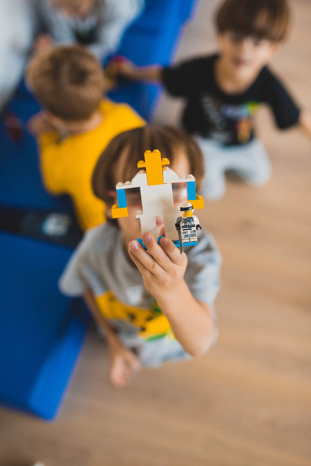 A child proudly displays a colorful Lego creation while three other children play on a wooden floor near blue furniture. Attention is focused on the Lego toy, with the blurry details of the children playing captured perfectly in this event photo. 