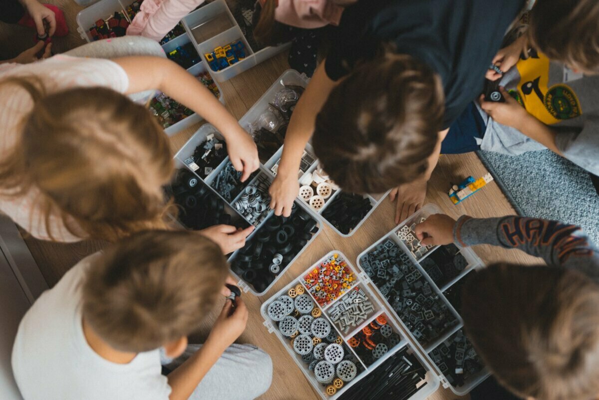 A group of children gather around a variety of containers filled with LEGO pieces, enthusiastically sorting and assembling them. The photo, a perfect example of event photography, is taken from above and shows their concentrated activity on the wooden floor. 