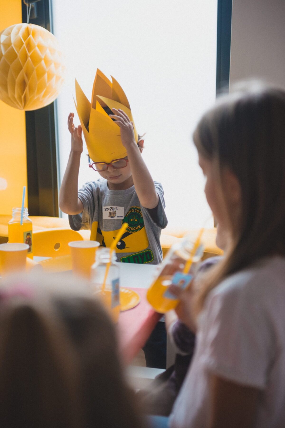 A child wearing a yellow paper crown corrects it while sitting at a table during the event. A child wearing glasses and a gray T-shirt is surrounded by other children holding yellow drinks with straws. The stage, ideal for event photography, is decorated with vibrant yellow and orange decorations.  