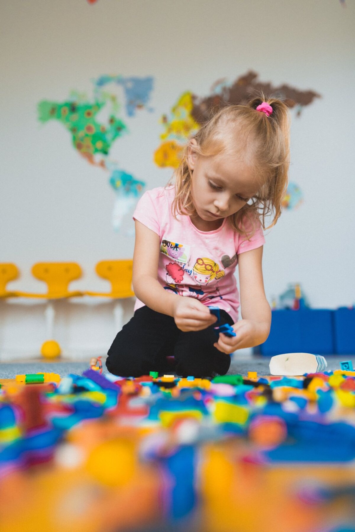 A young girl with blonde hair tied in a pink ponytail sits on the floor and is engrossed in playing with colorful blocks. She is dressed in a bright pink T-shirt with colorful graphics. In the background is a map of the world and yellow chairs that capture the charming moment of event photography.  