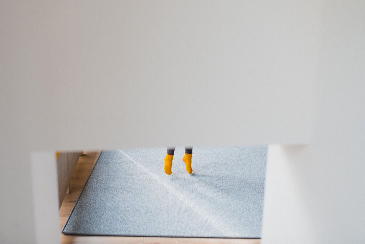 Minimalist photograph showing the partially visible legs of a child in bright yellow socks, jumping or standing on a light blue carpet, surrounded by white walls. The image captures a sense of movement and playfulness with the elegance often seen in event photographer Warsaw. 