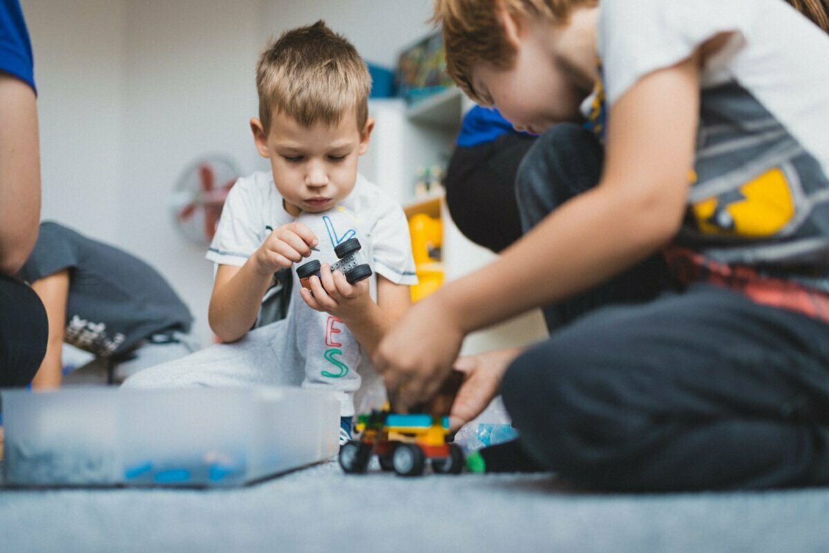 Two young boys are concentrating on assembling toy models from blocks. One boy, dressed in a white shirt, holds a piece and watches it carefully. The other boy, dressed in a gray shirt, kneels and works on a small toy vehicle from a nearby box of blocks - a cute moment perfect for event photography.  