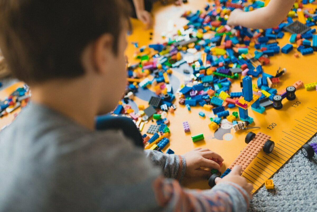 A child is playing with colorful LEGO bricks on a yellow mat, surrounded by various elements and minifigures. The child's hands are assembling the piece, while other children can be seen in the background, also busy with LEGO bricks - a perfect scene for event photography showing the joyful moments. 