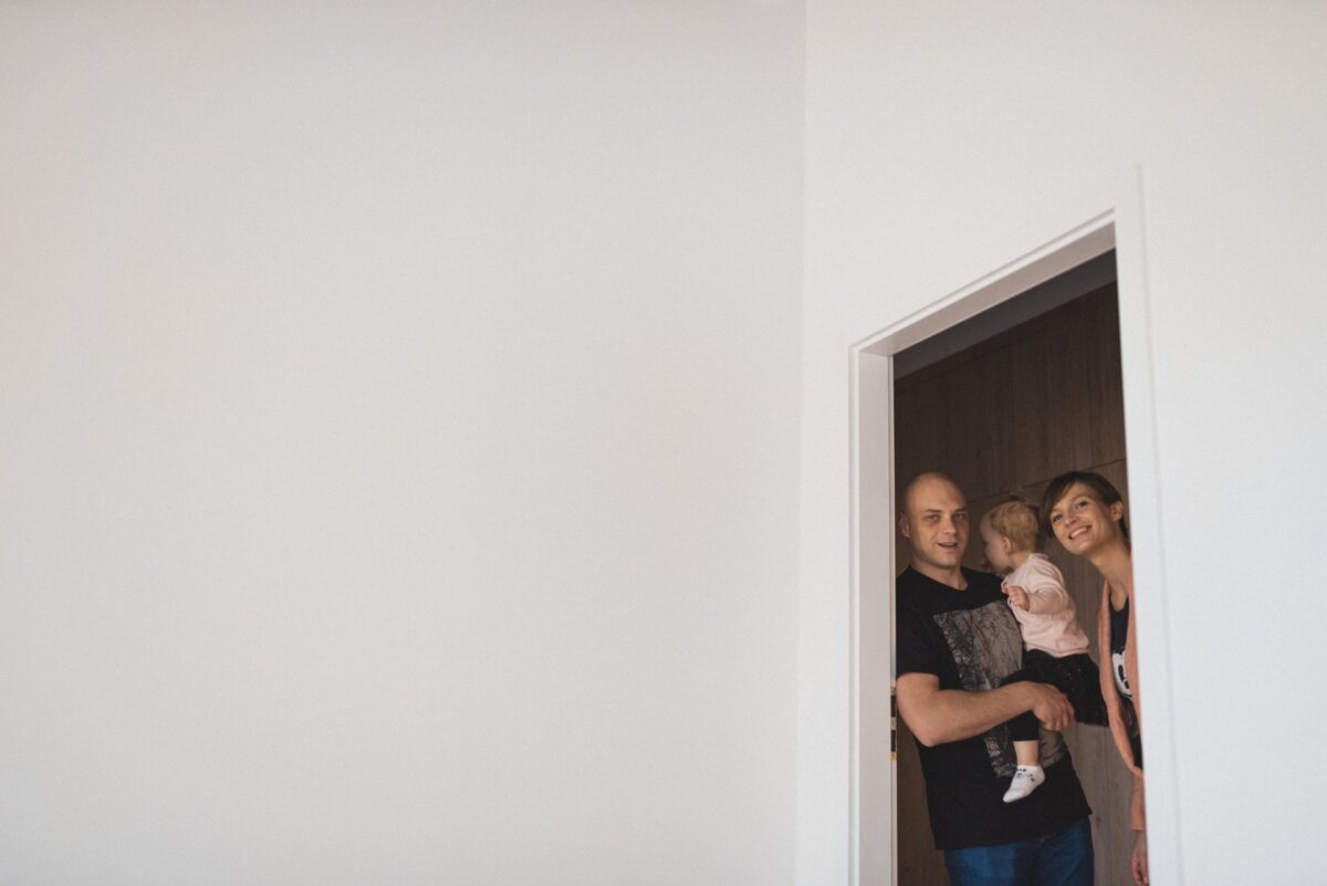 In the doorway stand a smiling man and woman, holding a small child, radiating happiness and closeness. A backdrop of a simple white wall with minimalist decor highlights the bright, intimate setting. Perfect for event photography, capturing such authentic moments is what makes event photojournalism so special.  