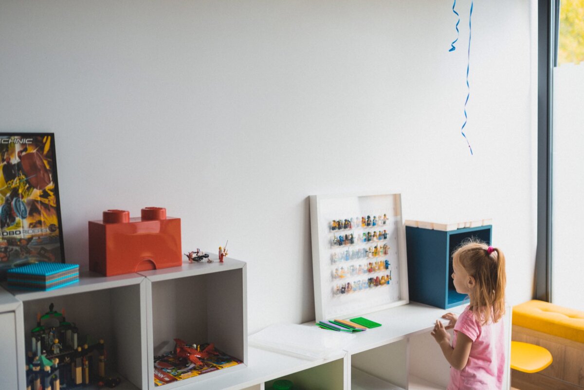 A young girl with a ponytail, wearing a pink shirt, stands in a playroom looking at a framed Lego set set on a white shelf. The room, like an event photographer warsaw, has white shelves filled with toys, including a large red Lego storage box and toy cars against a plain white wall. 