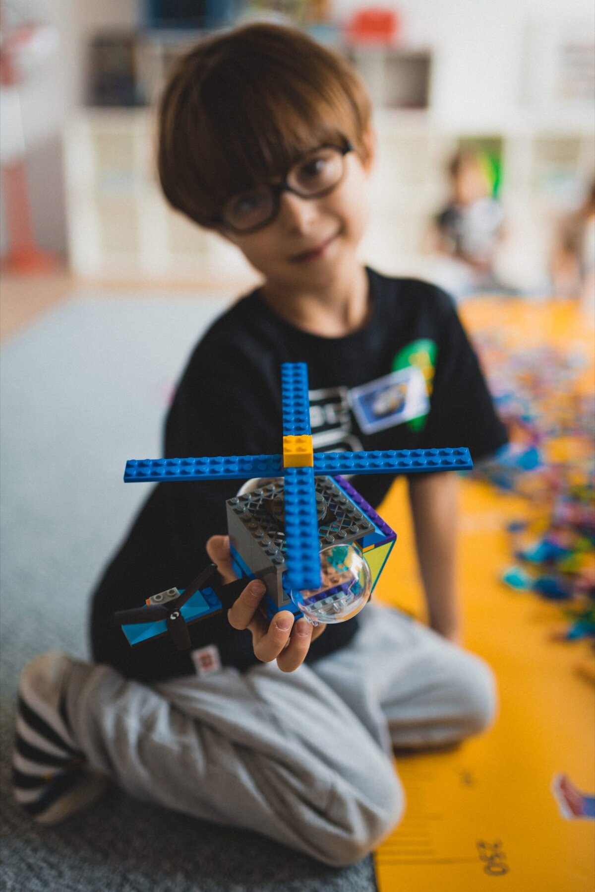 A young child wearing glasses is holding a blue and yellow Lego piece resembling a small helicopter or windmill. The background is a bit blurry, but shows a game area with more Lego pieces scattered on the floor. The child is smiling, making it perfect for event photography, capturing happy moments.  