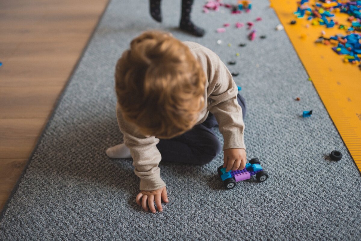 A child with light brown hair sits on a gray carpet and plays with a small blue and purple car. Multicolored blocks are scattered around, and a yellow mat is partially visible on the right side. In the background you can see the legs of another child, making it the perfect moment for event photography.  