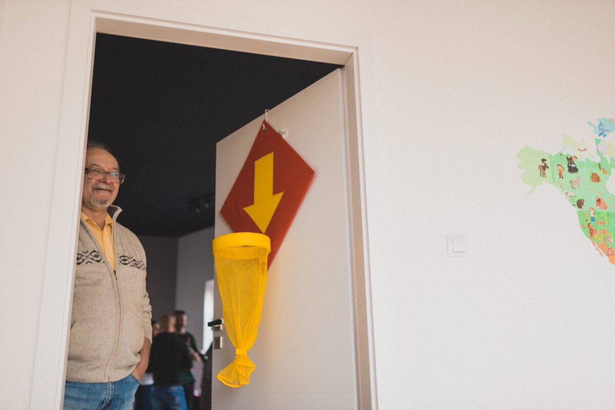 In the doorway stands a smiling man with glasses, wearing a beige patterned sweater. Above him hangs a red sign with a large yellow arrow pointing downward. In the background, a section of a colorful map can be seen on the right wall - a scene ideal for photo coverage of events.  