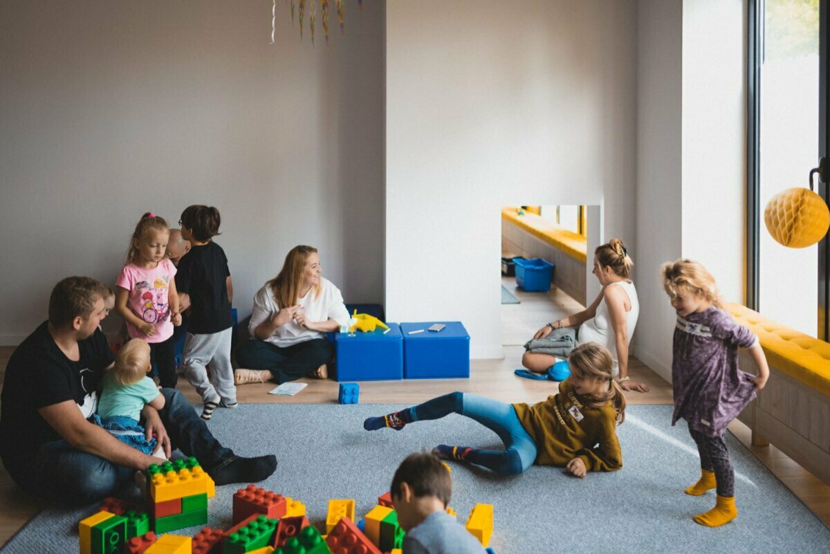 A spacious, light-filled room where children and adults engage in various activities. Some children play with large blocks on the floor, while others interact or relax. Adults are present, sitting or supervising in a casual, relaxed atmosphere - perfect for event photography.  