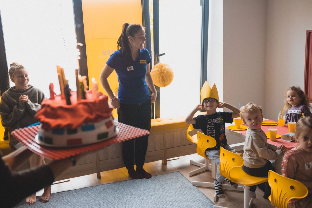 A group of children is celebrating a birthday at a party. An adult holds a colorful cake with candles while another adult stands nearby. A child wearing a crown looks excited. Other children sit at a table with party hats and cups. The room is well-lit and festive, perfect for photo opportunities of the events.    