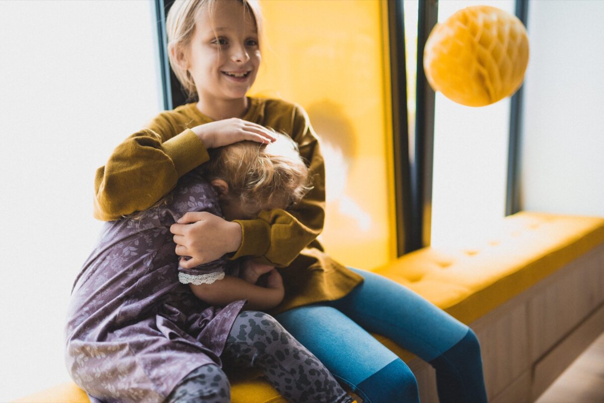 Two children are sitting on a yellow bench by the window. The older child, dressed in a mustard-colored sweater, smiles, gently holding and comforting the younger child, who is dressed in a purple dress and puts her face in the older child's lap. A yellow pendant completes the offering. photo coverage of the events.   