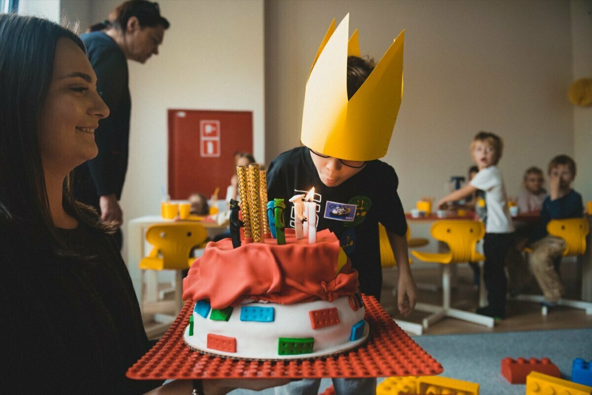 A child wearing a paper crown leans over to blow out the candles on a colorful LEGO-themed birthday cake held by a smiling adult. Other children sit at a table in the background and watch the events unfold - a perfect moment for event photography. The venue looks like a lively party room.  