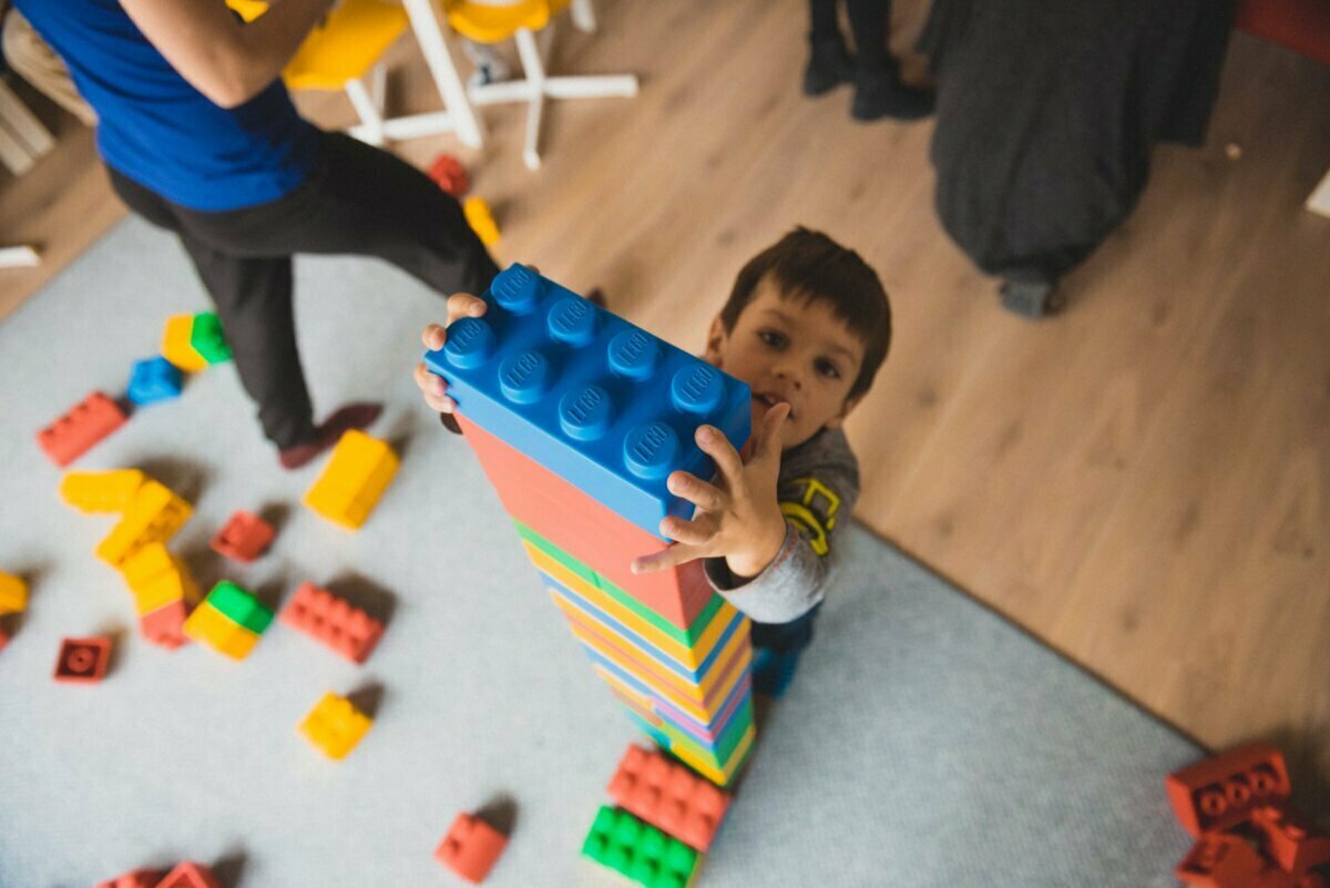 A child, standing on a carpeted floor, arranges large, colorful blocks into a tall tower. Other blocks are scattered around. An adult can be partially seen in the background, capturing the moment for an event photo shoot.  