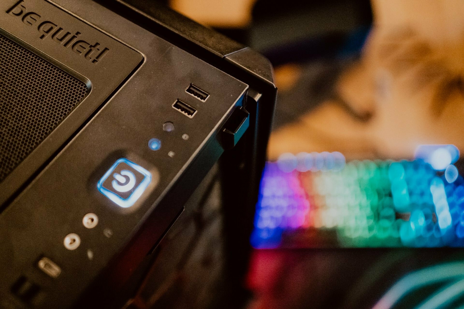 Close-up of the top of a black computer tower with multiple USB ports, a blue backlit power button and the "be Quiet!" logo. A colorful RGB backlit keyboard with fuzzy multicolored lights is partially visible in the background during the press briefing.