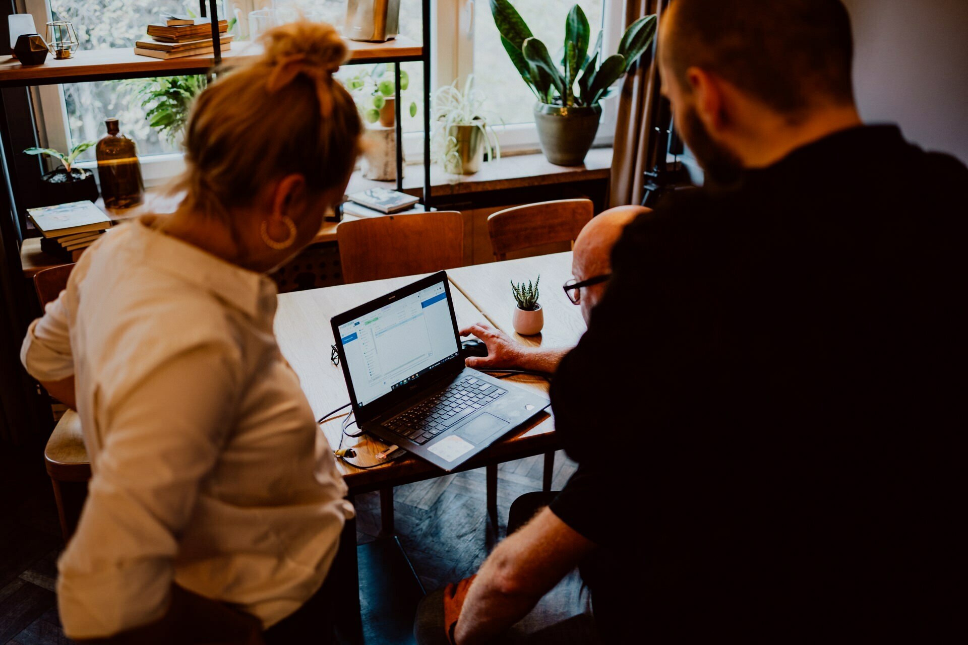 Three people are gathered around a table, focused on a laptop screen displaying a document, perhaps about an upcoming press meeting. The environment appears to be an informal meeting space with wooden furniture and potted plants on shelves and windowsills. One person points to the laptop screen.  