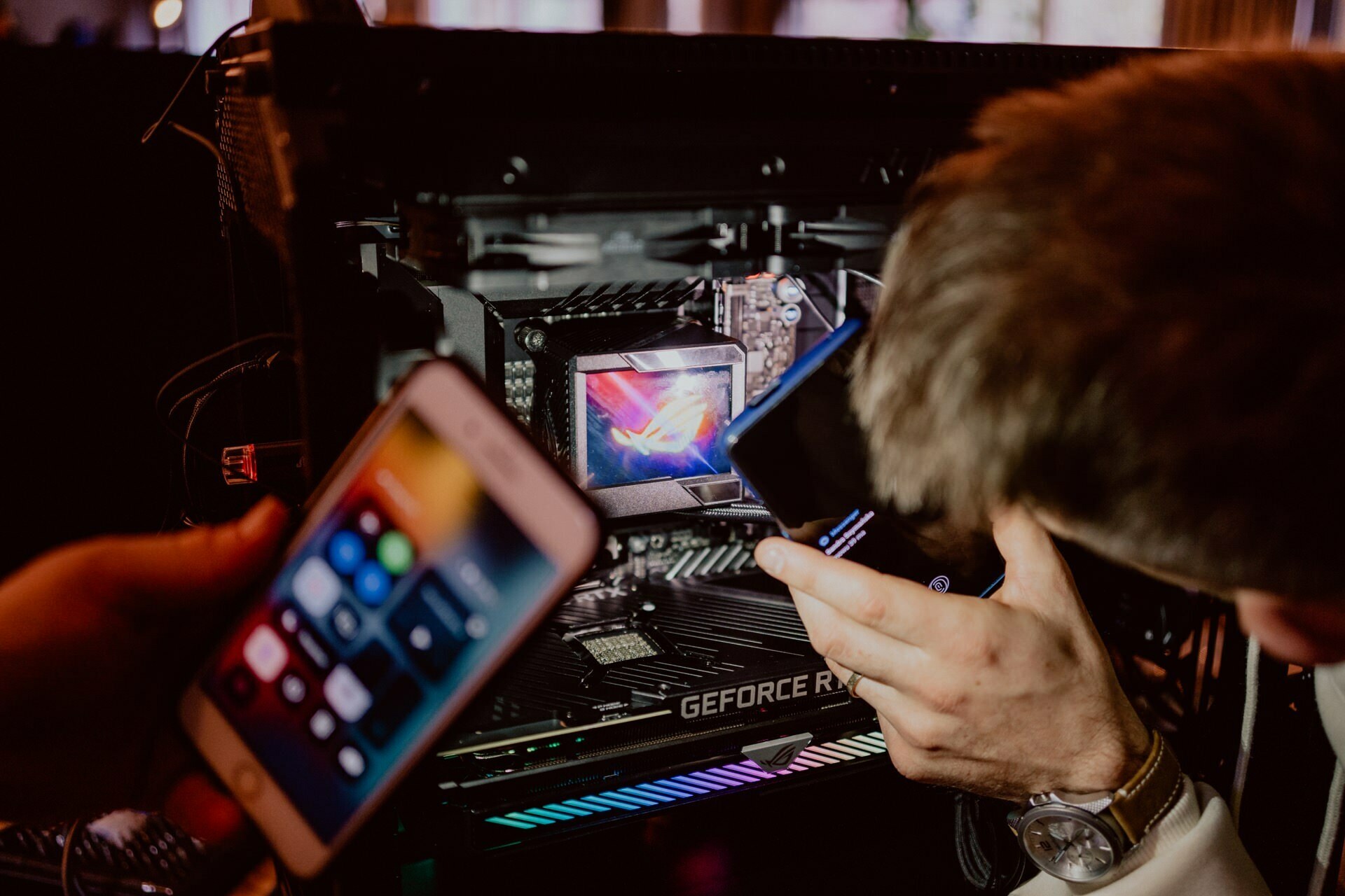 Two people examine the inner workings of a desktop computer. One person is holding a smartphone with a visible screen, while the other person appears to be using another smartphone as a light source. A GeForce RTX graphics card is visible inside the computer, which brings to mind technology enthusiasts at a press briefing.  