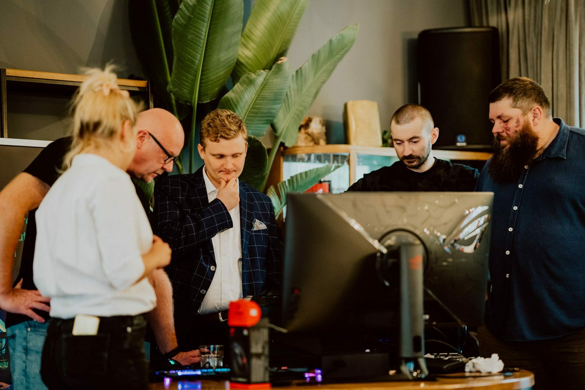 A group of five people, four men and one woman, are gathered around a large computer monitor. They are engaged in a discussion, appearing focused and thoughtful. There is a large plant in the background and various objects scattered on the desk, creating an atmosphere reminiscent of a press meeting.  