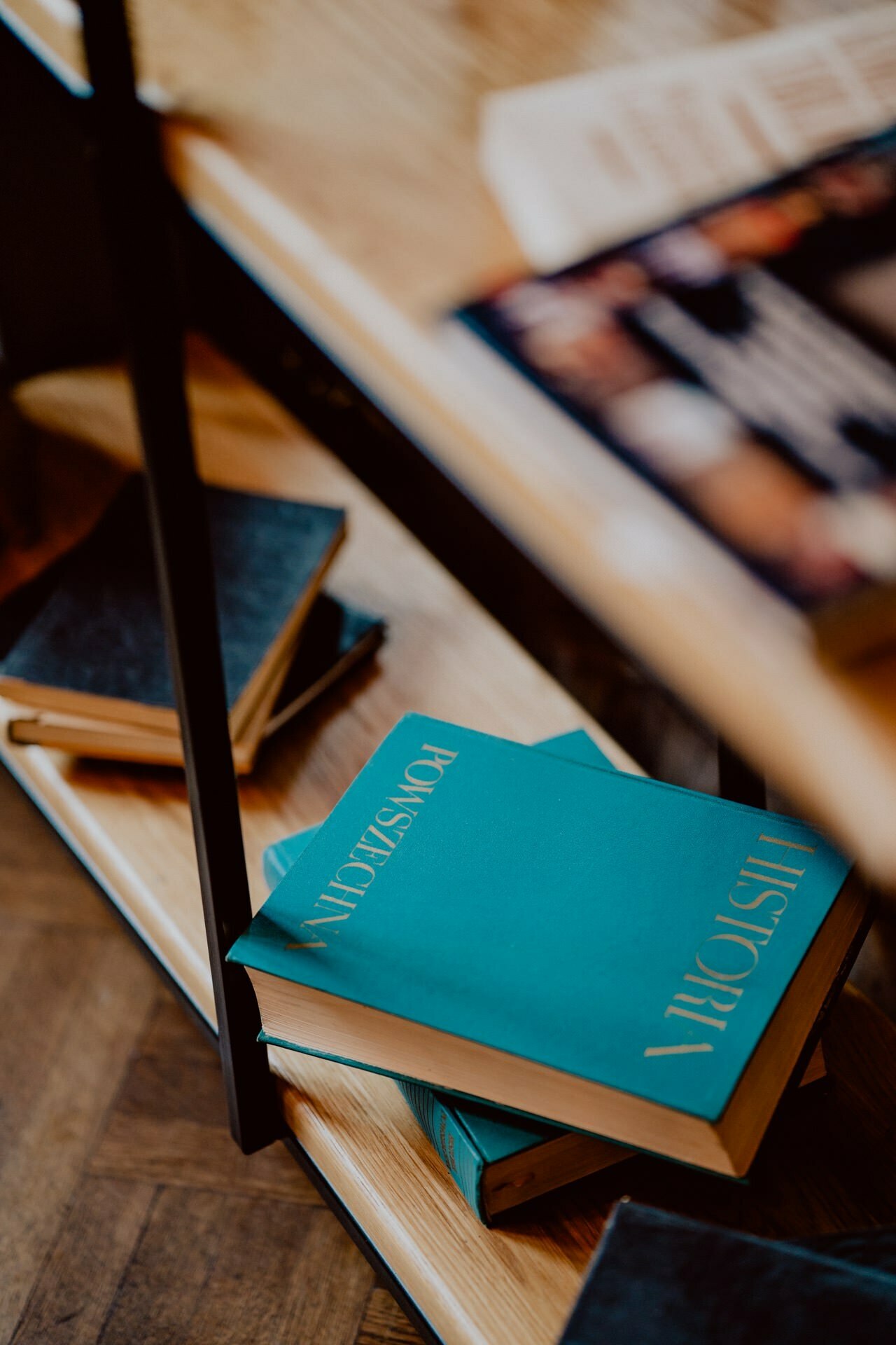 A bookshelf on which several books are scattered, including a clear turquoise-colored book placed at an angle on the lower shelf. Other books with darker covers are also visible on the shelf and in the background, which is reminiscent of a photo report of events. The floor is wooden.  