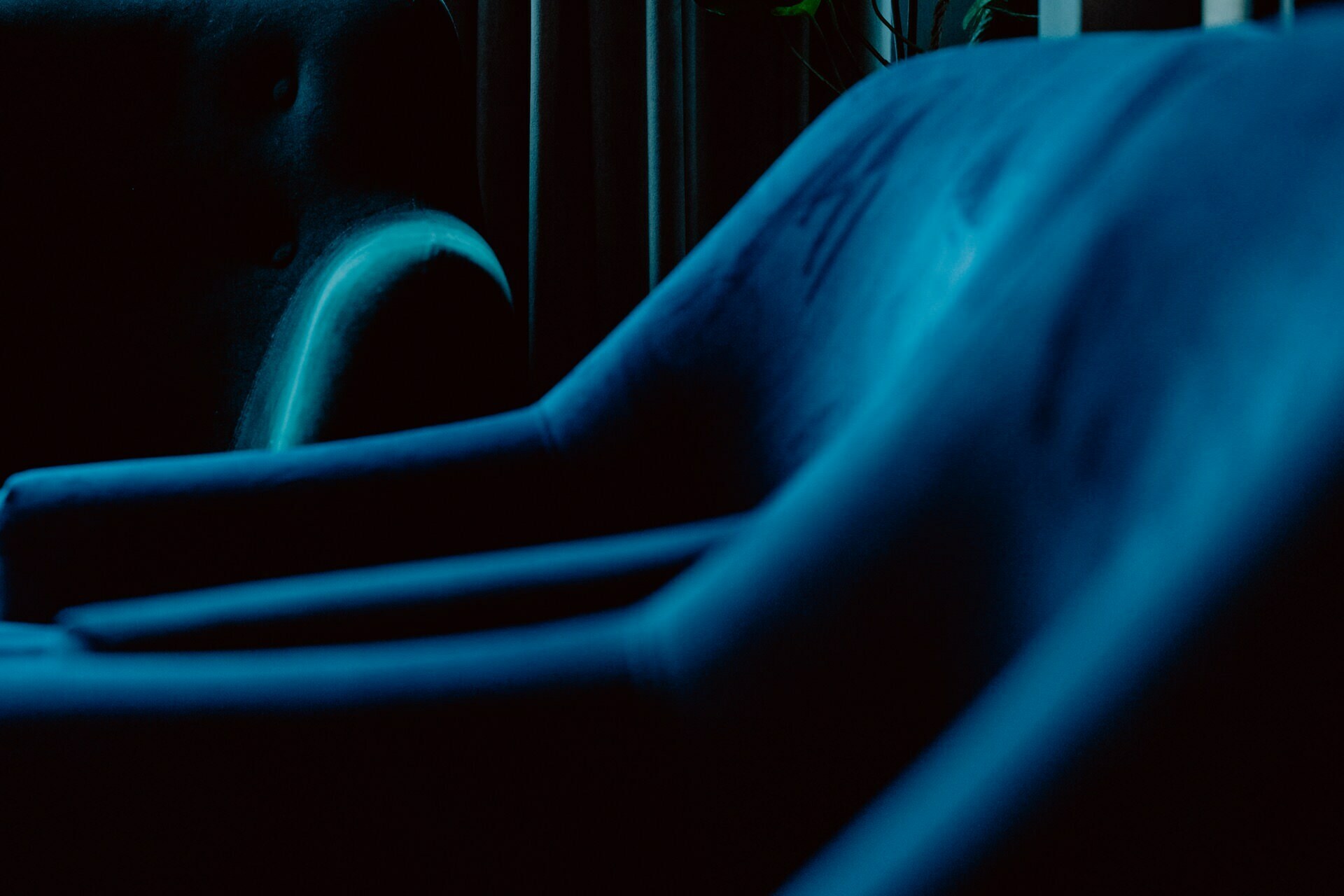 Close-up of a set of modern dark blue upholstered chairs in low light. The chairs, perhaps prepared for a press meeting, are lined up and lean to the right, with smooth curves and soft textures highlighted by subtle lighting. An indistinct, dark background with curtains.  