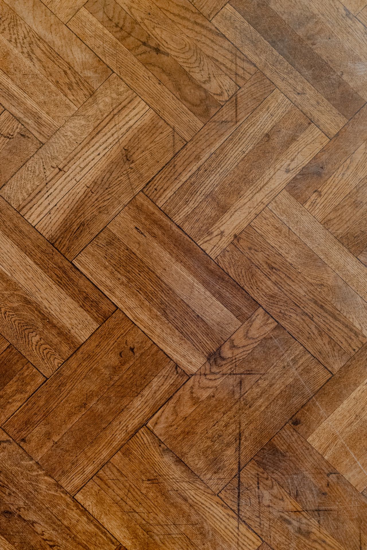 Close-up of a wooden parquet laid in a herringbone pattern, reminiscent of an elegant press briefing. The natural grain of the wood and differences in shades create a textured and intricate visual effect. The surface shows minor signs of use and scratches, which add character to the floor.  