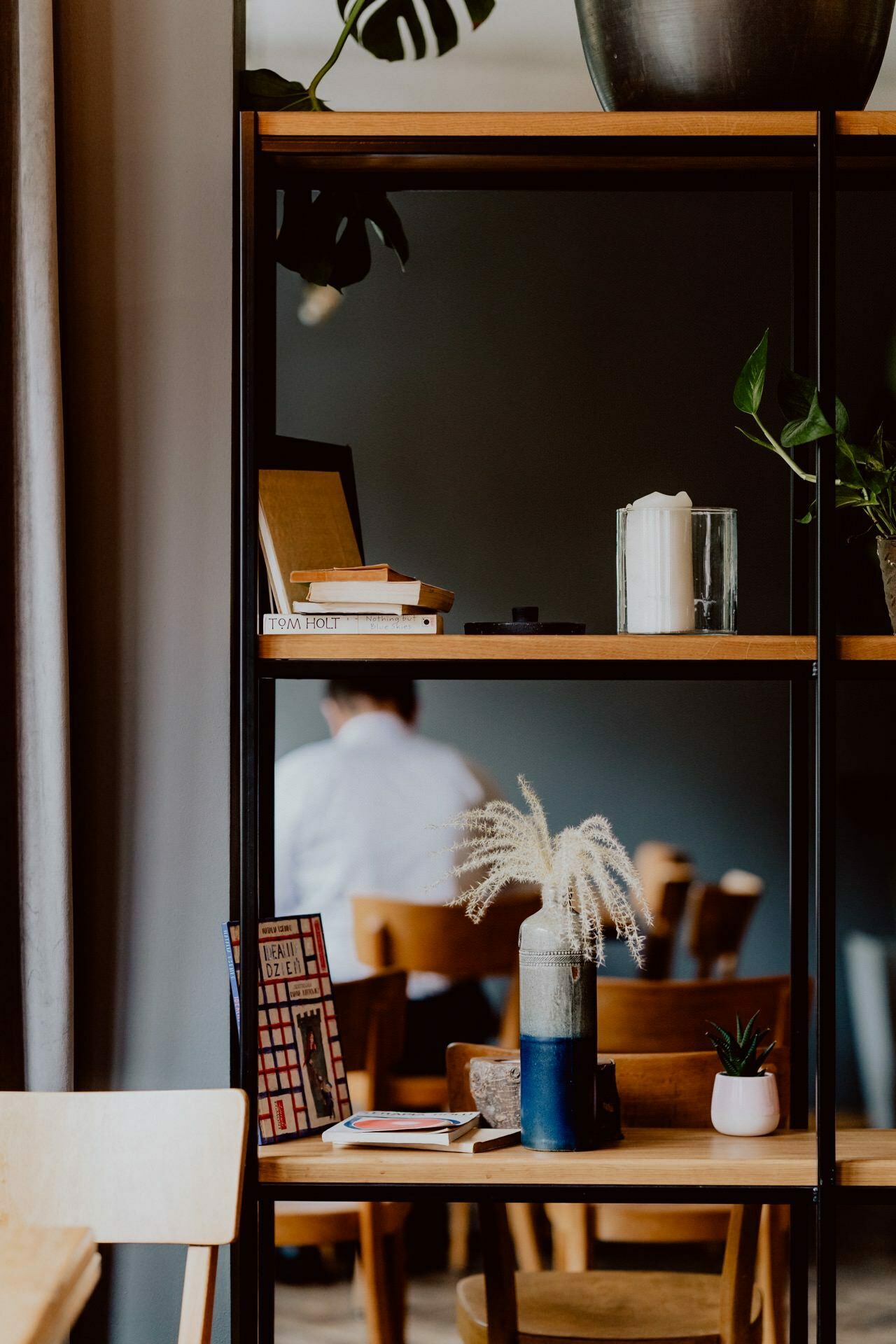 The modern living space features wooden and black metal shelves that hold decorative items such as books, candles and potted plants. In the background, a person sits on a wooden chair at a table during a press meeting. The room has a minimalist design with natural light.  