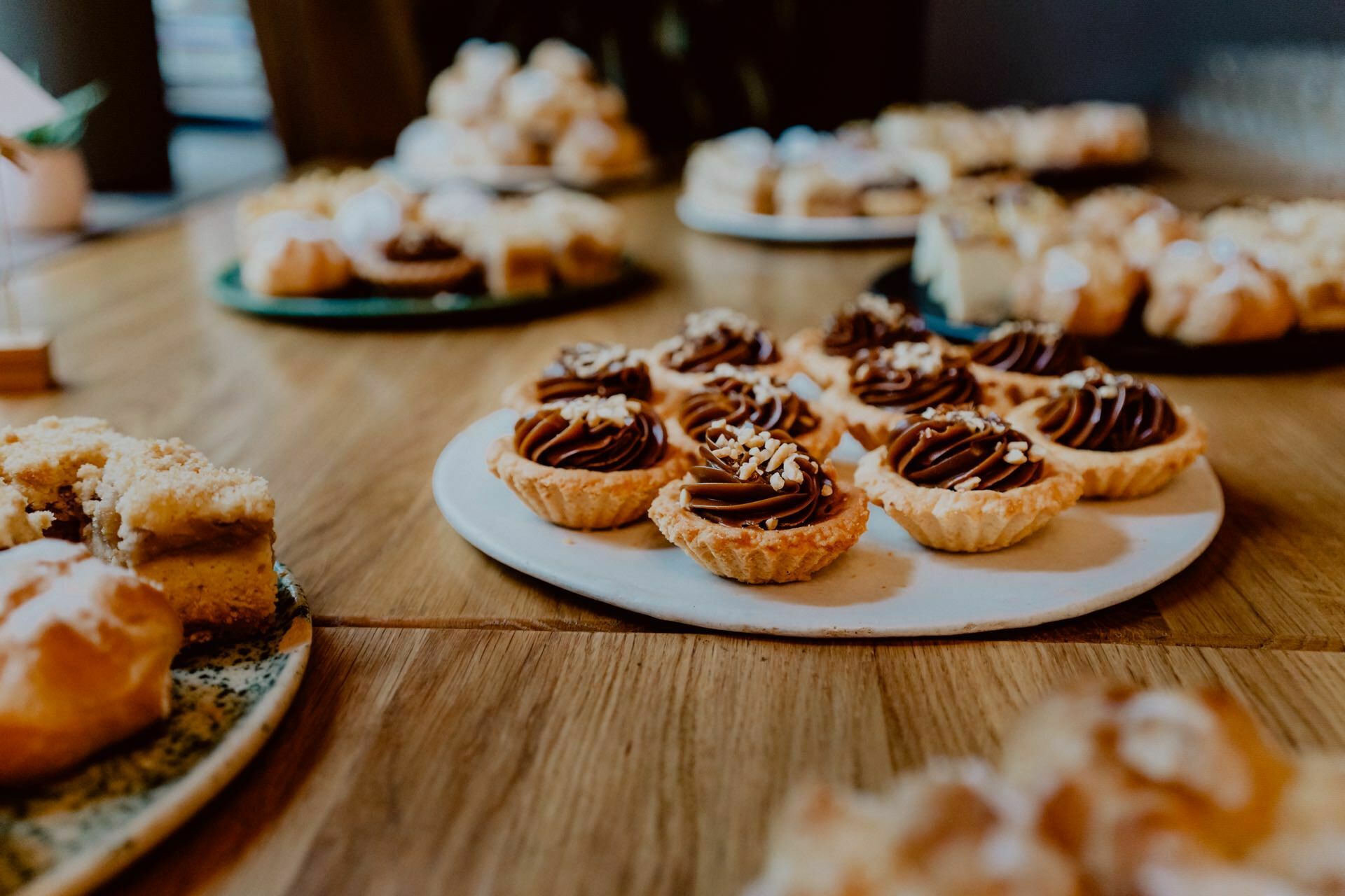 The table is set with a variety of desserts, including small tartlets sprinkled with chocolate chunks and chopped nuts on a white plate, and other cakes and pastries on various plates in the background. The desserts are displayed in a warmly lit room, ideal for an elegant press meeting. 