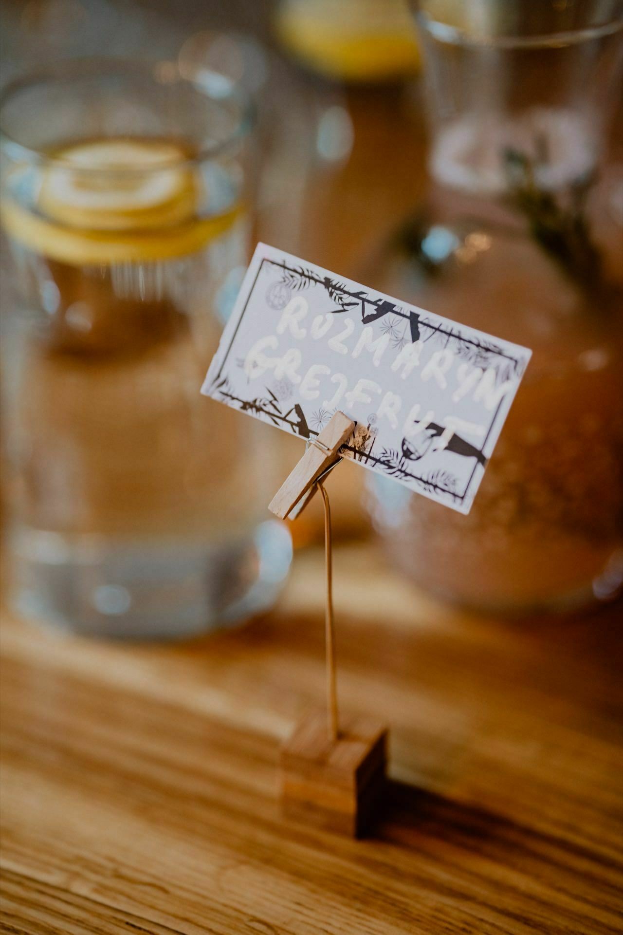 A business card held on a stand with a wire clip is placed on a wooden table. Behind it are glasses of water with lemon slices and a glass jar with a brown drink. The card, probably from a press meeting, has a floral motif with illegible text.  