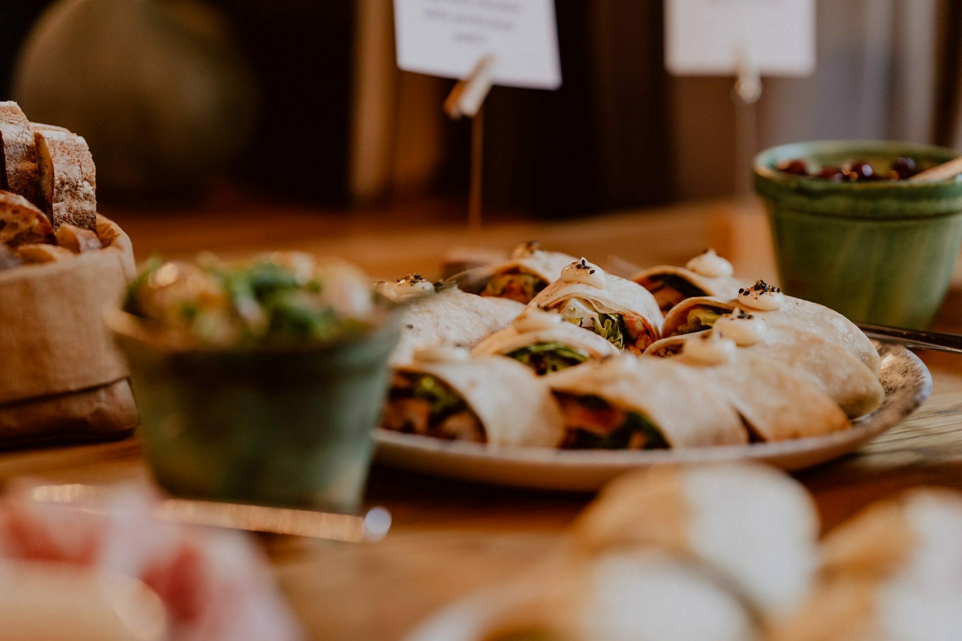 Close-up of a table with various food products at a press meeting. In the foreground is a plate of sliced wraps filled with greens and other ingredients. On the left a basket of sliced bread, on the right two green ceramic salad bowls.  