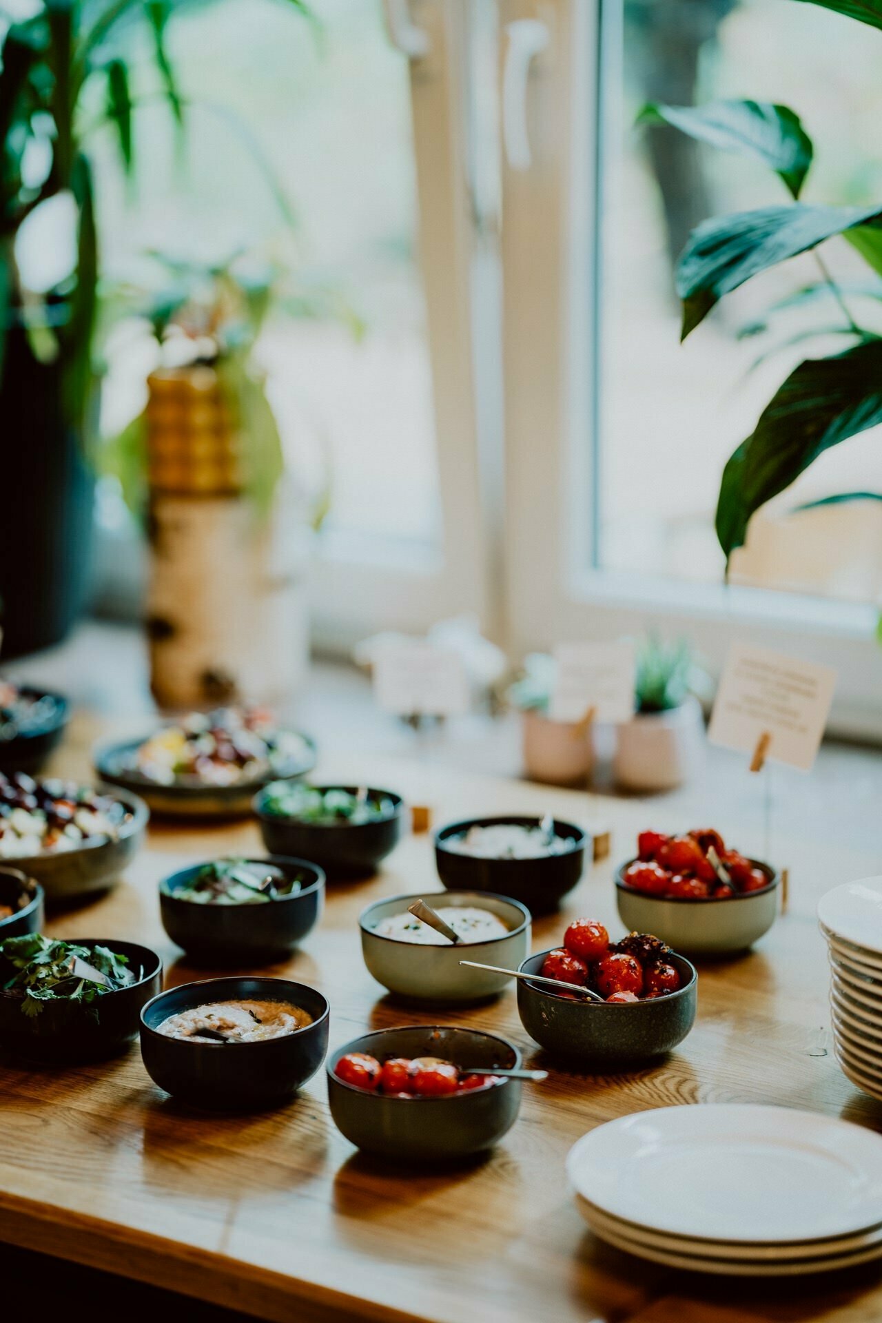 The wooden table is set with various small bowls containing salads, roasted vegetables and other colorful dishes. In the background, plants by the window create a fresh and inviting atmosphere, perfect for an intimate press meeting. 