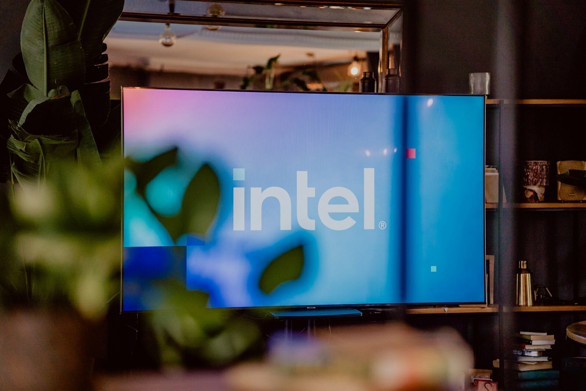 The TV screen displaying the Intel logo on a blue background is sharp, with blurry green plants in the foreground. Placed in a modern, stylish room decorated with various design elements, the TV suggests it could be set up for a press meeting. 