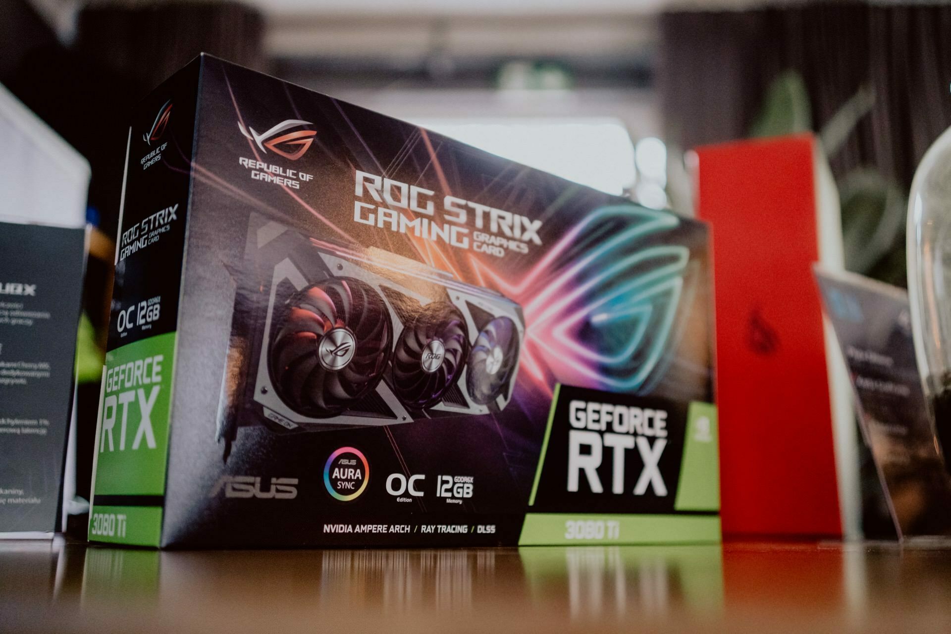 A close-up of the ASUS ROG Strix Gaming graphics card box with "GeForce RTX" and "12GB GDDR6X" printed on it, presumably presented at a press briefing. The box contains the graphics card with three cooling fans and RGB lighting effects, while various other components and packaging can be seen in the background. 