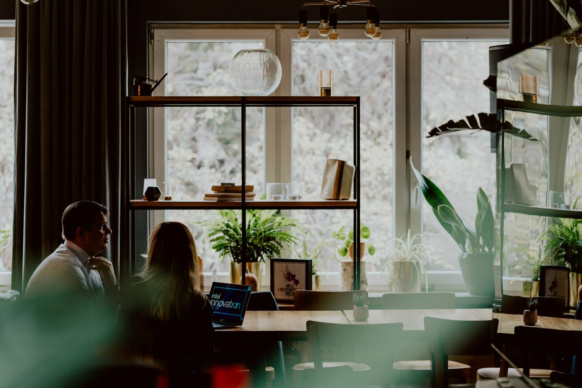 A well-lit room where two people sit at a table, facing a large window with shelves containing books and plants. One person is using a laptop, probably preparing for a press meeting. There are more plants near the window, and the scene looks cozy and peaceful.  