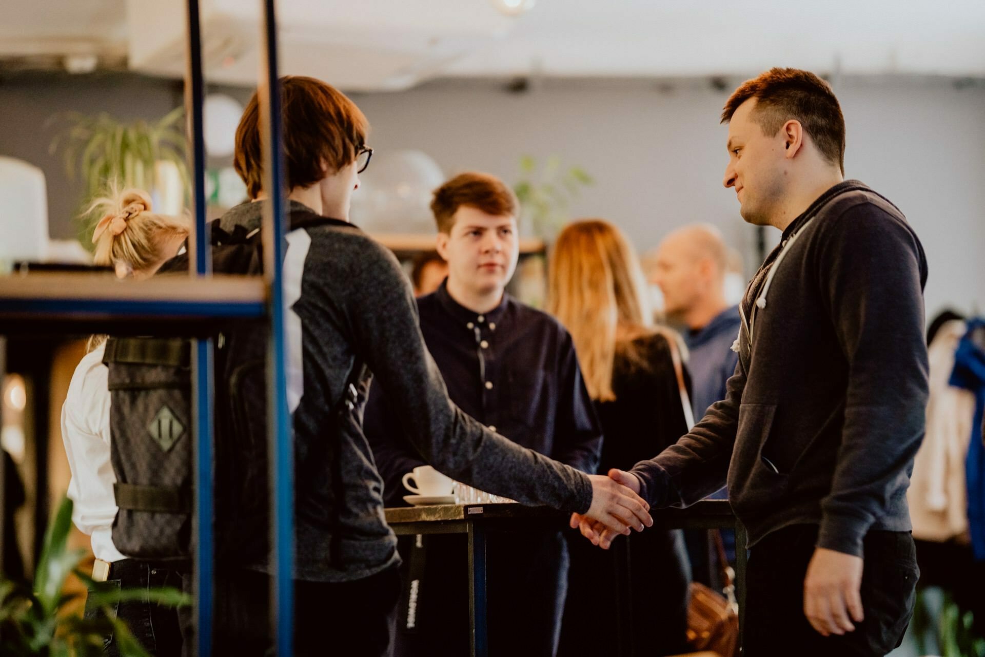 A group of people having a conversation in a seemingly casual room. Two people in the foreground are shaking hands, while others are standing and talking in the background. The atmosphere seems informal and friendly, reminiscent of a casual press meeting.  