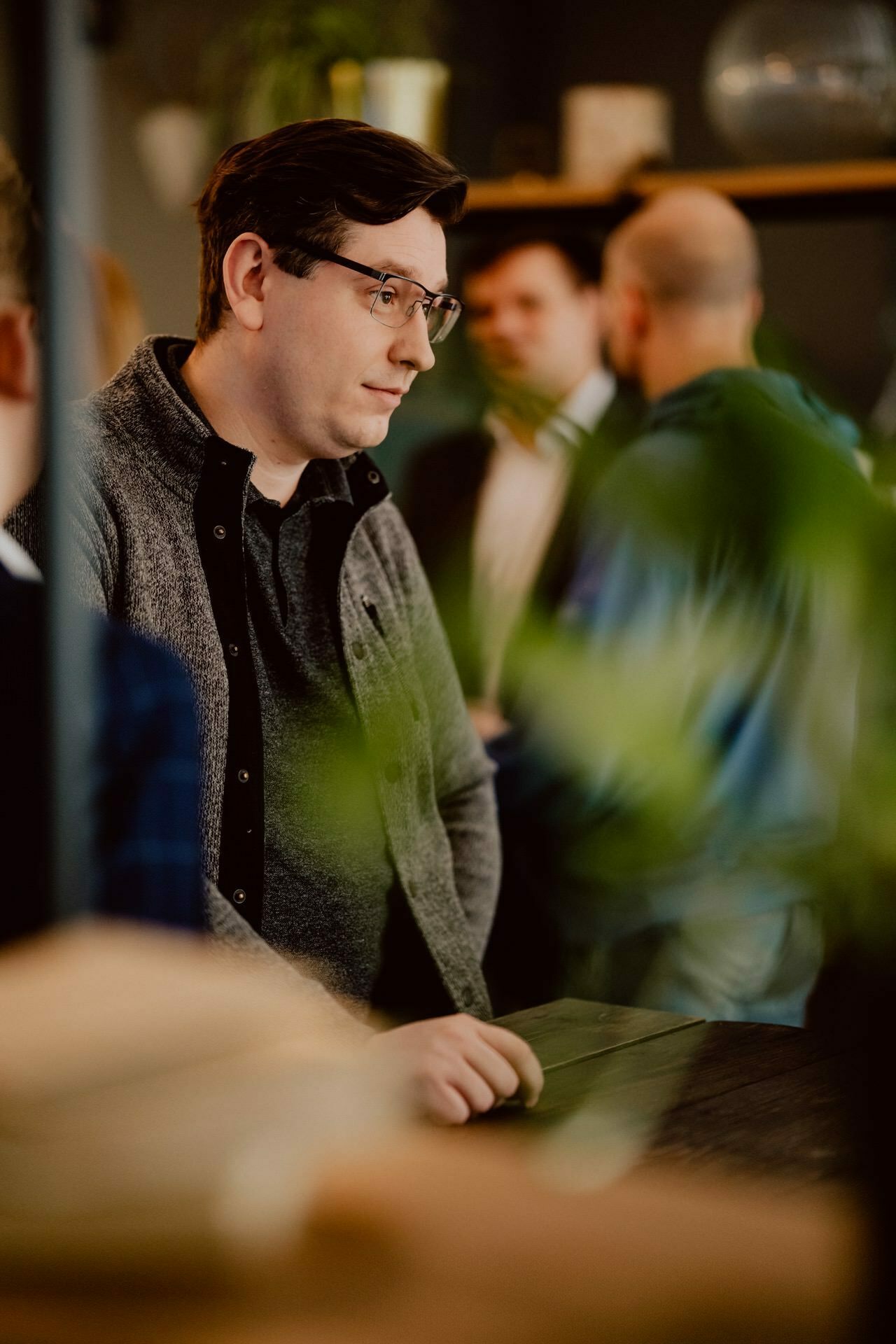 During a press briefing, a man with short, dark hair and glasses, wearing a black shirt and gray cardigan, stands in a room with his hands resting on a table. Other people and houseplants can be seen in the blurred background. 