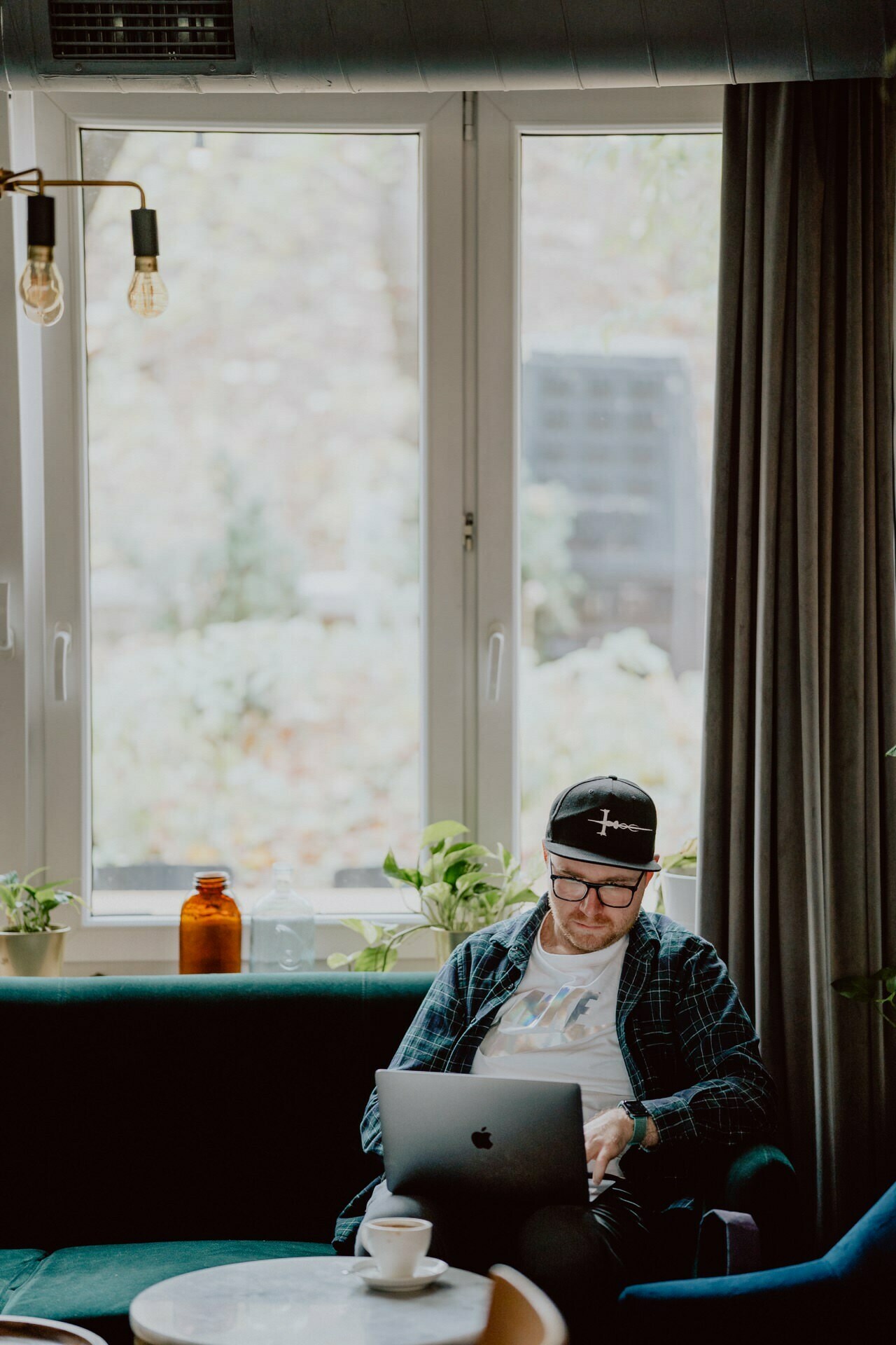 A man wearing a cap and glasses sits on a green couch with a laptop on his lap in a cozy room and prepares for a press meeting. A cup of coffee sits on a table in front of him, with large windows behind him letting in natural light. Potted plants are placed on the window sill.  