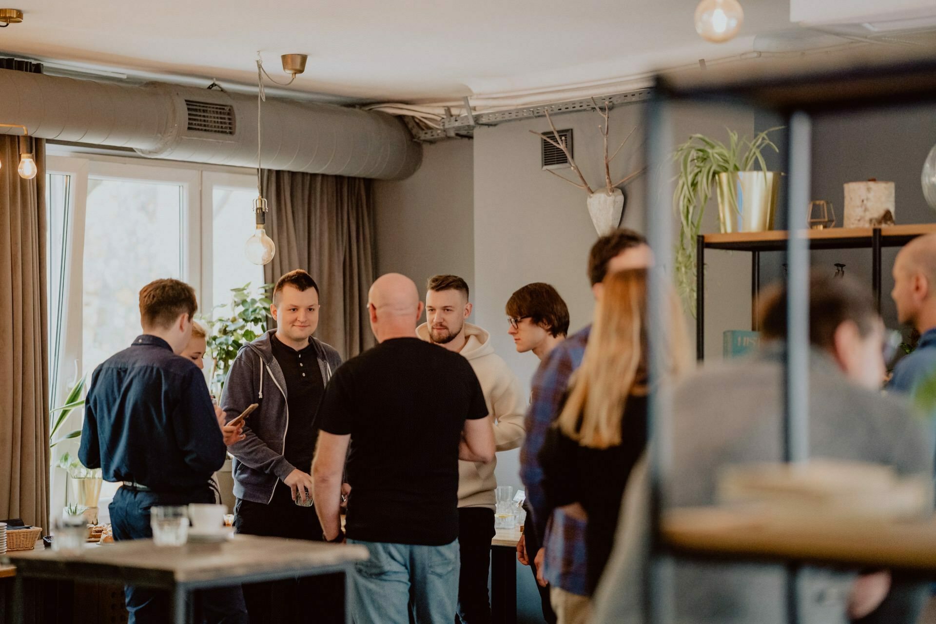 A group of people stand together and talk in a warmly lit, modern room. There are plants and shelves in the background, and large windows let in natural light. The participants, dressed in casual clothes and engrossed in conversation, seem to be enjoying an informal press meeting.  