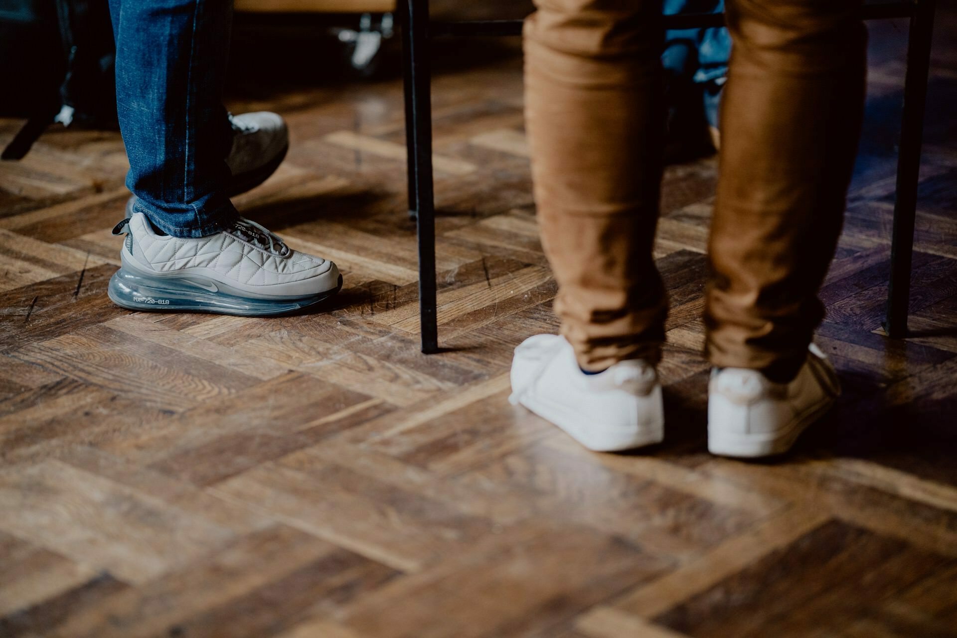 Close-up of two people standing on a herringbone wooden floor at a press meeting. One person is wearing blue jeans and gray tennis shoes, the other is sporting brown pants and white tennis shoes. Both are standing near a black metal table or chair leg.  