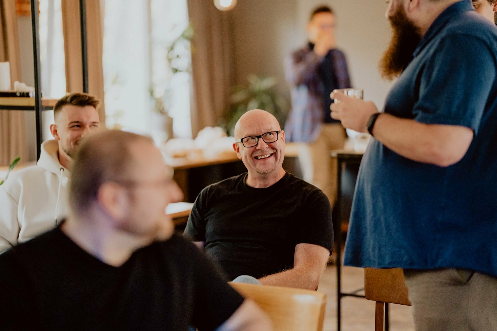 A group of people meet in a closed room. A bald man in glasses and a black shirt smiles and sits. Another man in a blue shirt stands holding a drink, presumably at a press meeting. Blurred figures can be seen in the background, creating a casual and friendly atmosphere.   