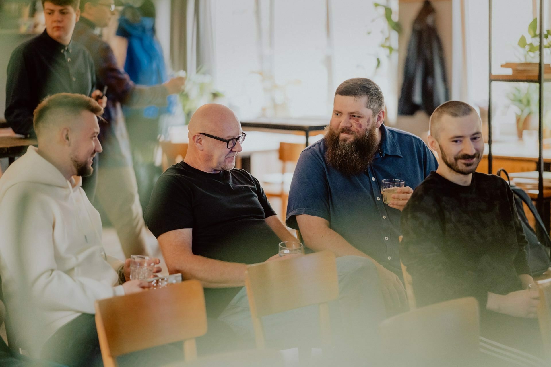 Four men sit together in a bright room, talking and laughing. Two hold drinks; one with a beard smiles broadly at the other man with glasses. The background depicts blurred figures and plants, creating a warm and relaxed atmosphere similar to a casual press meeting.  