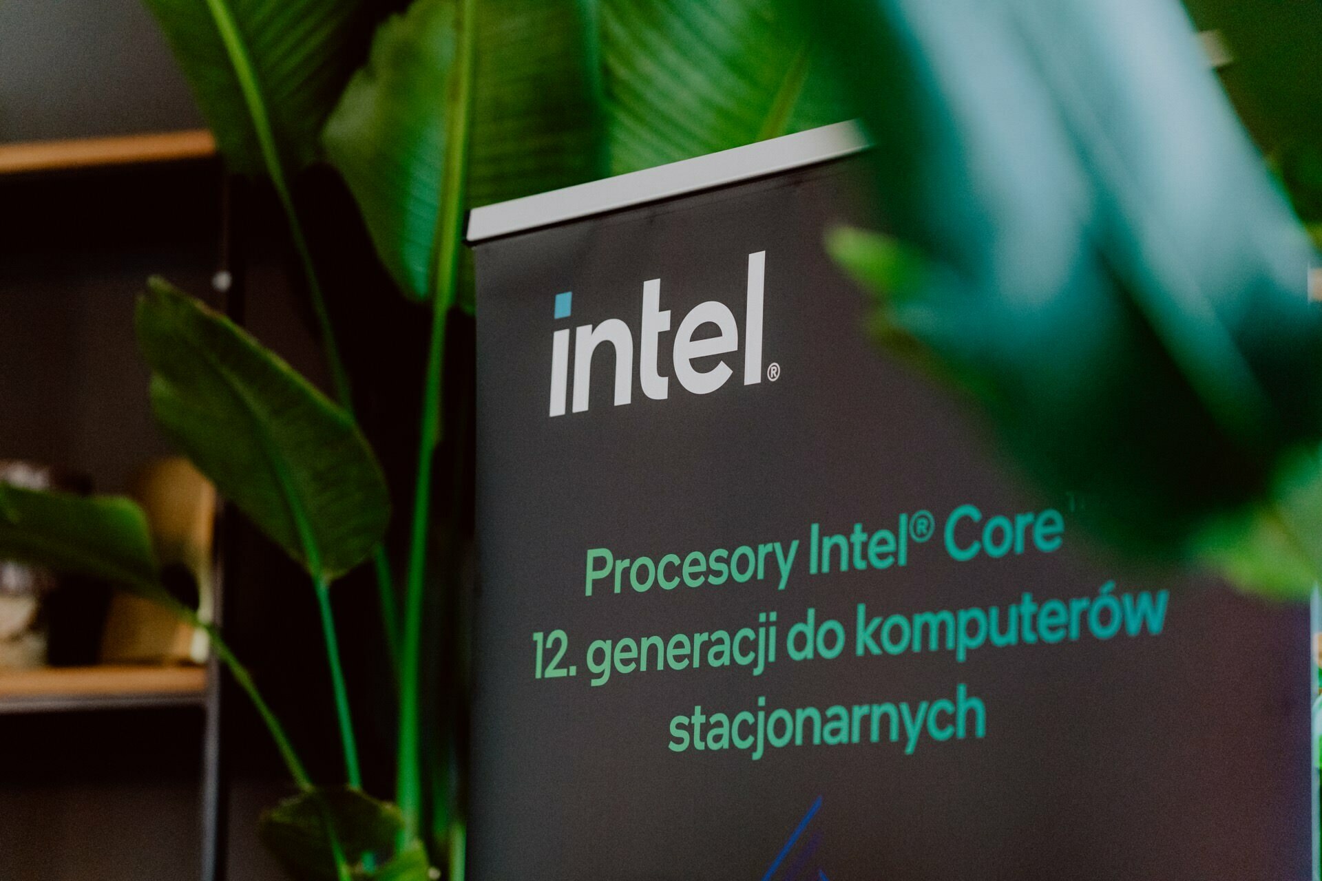 A banner with the Intel logo and text in Polish promoting Intel's 12th-generation Core desktop processors is surrounded by green plants. Ideal for event photo coverage or any event photography, this set seamlessly blends technological innovation with natural aesthetics.  