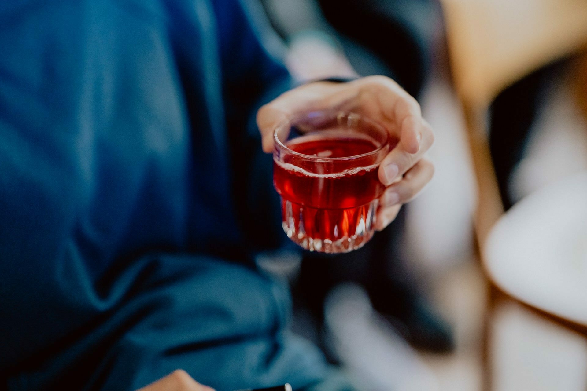 A person in a blue shirt holds a glass of red liquid, probably a drink, in his hand. The image is closely cropped, focusing on the hand and the glass. The background is blurred, highlighting the glass and the drink - perhaps capturing a moment at an informal press meeting.  