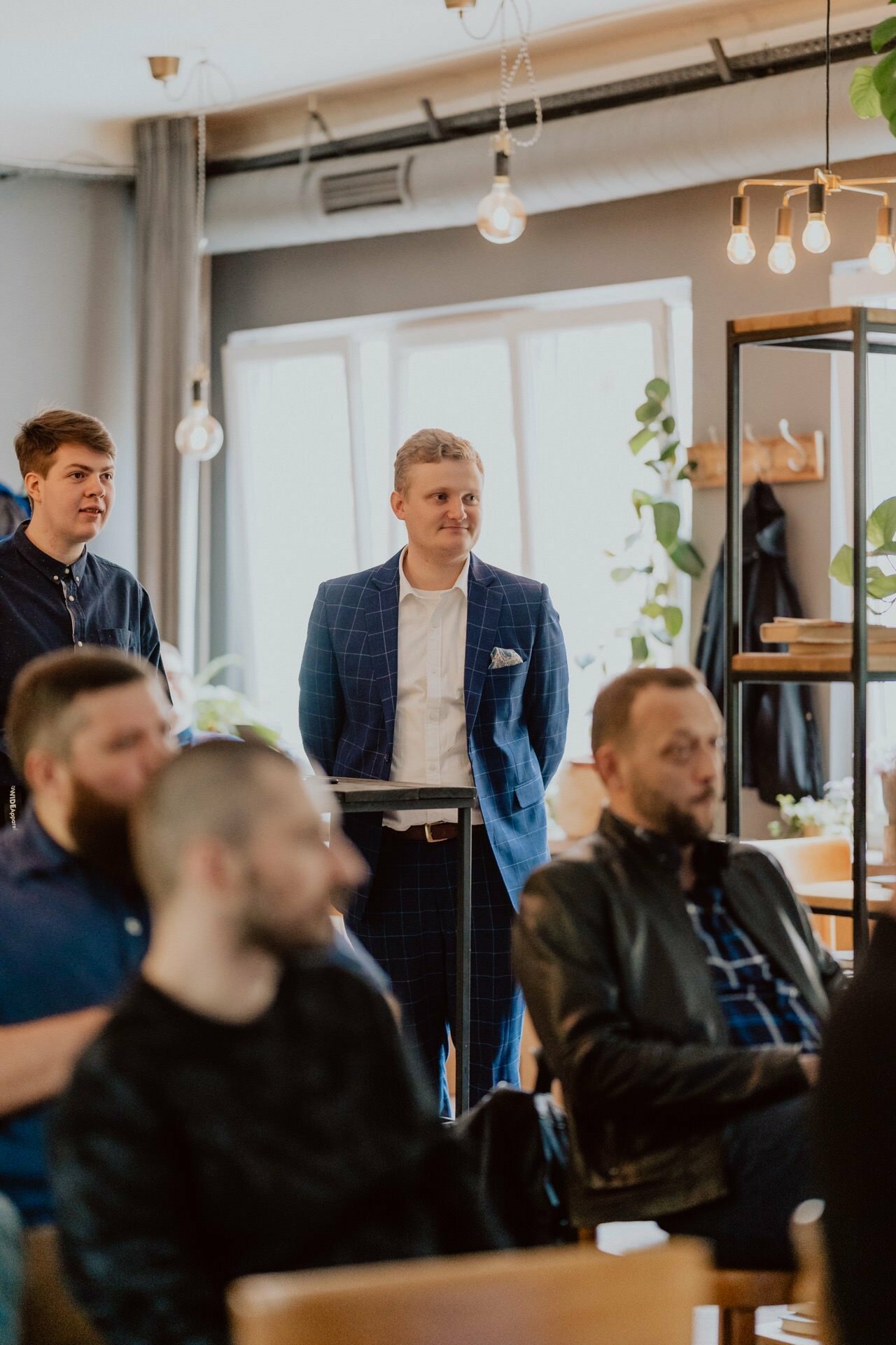 Next to the podium stands a smiling man in a blue plaid suit, and next to him is another man in a dark shirt. Several seated people in casual attire stand in the foreground and listen attentively. The room is well-lit, with large windows and modern decor befitting the scene of a press meeting.  