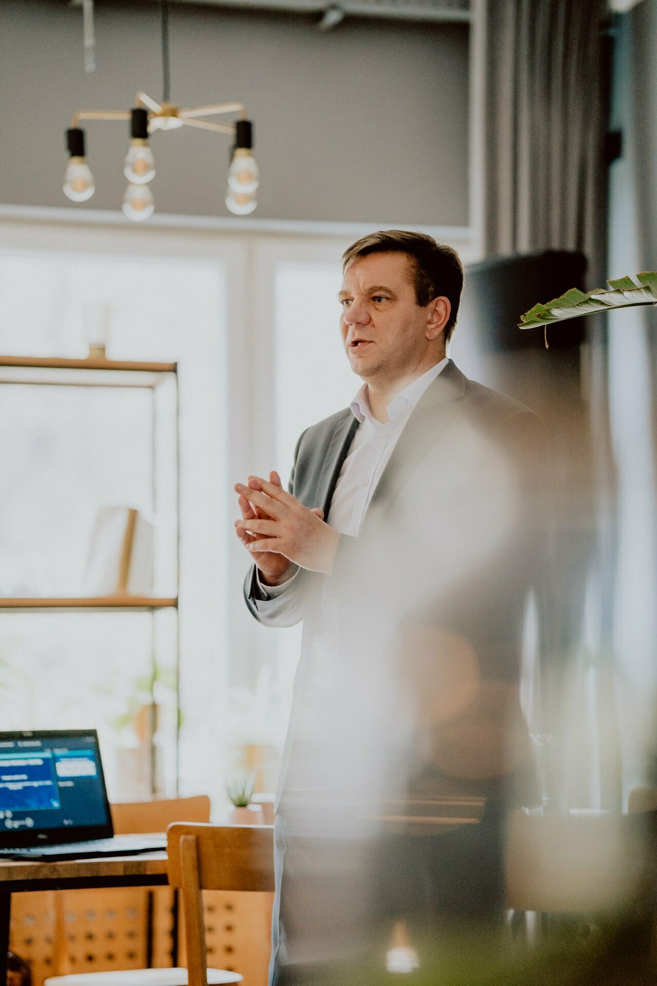 A man in a suit stands and speaks in a bright room with large windows, seemingly hosting a press meeting. A laptop computer with an app open is visible on a wooden table in the foreground. The room also has a bookshelf, and modern pendant lights hang from the ceiling.  