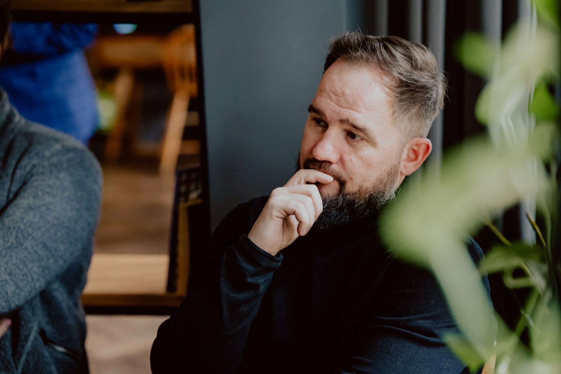 A bearded man wearing a long-sleeved black shirt sits and appears to be deep in thought, with his hand resting on his chin during what appears to be a press meeting. The background is slightly blurred, showing some furniture and greenery. 