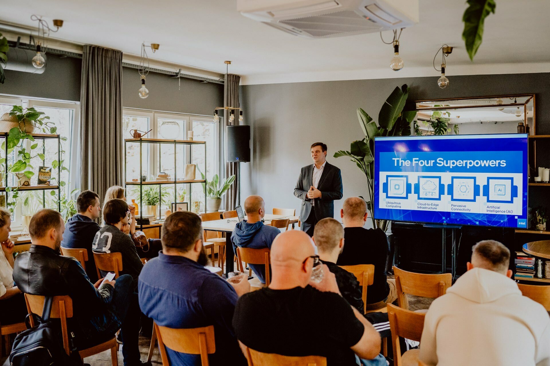 A man in a suit stands in front of a group of seated people and gives a presentation. Behind him, a large screen titled "Four Superpowers" displays diagrams. The room, reminiscent of a professional press meeting, is well lit with natural light, decorated with plants and modern furniture.  