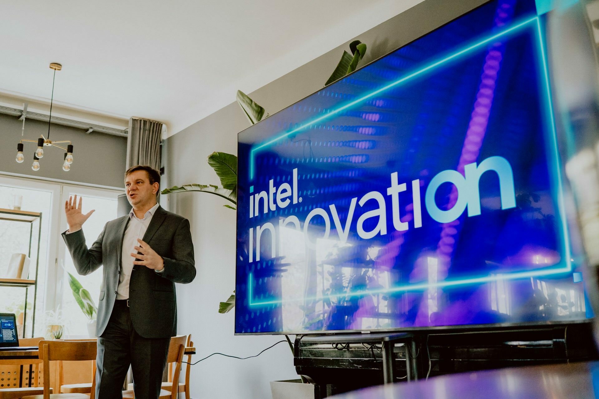 A man in a suit is giving a presentation in a modern room with Scandinavian-style decor. He stands next to a large screen that displays the words "Intel Innovation" on a vivid, technology-inspired background. During the press meeting, natural light streams in through the windows.  